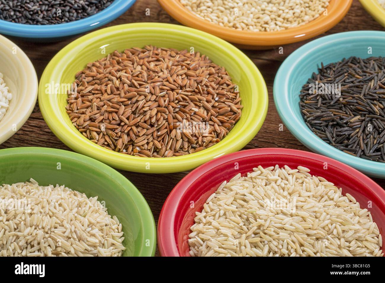 Una varietà di grani di riso su ciotole di ceramica colorata contro un legno a grana Foto Stock
