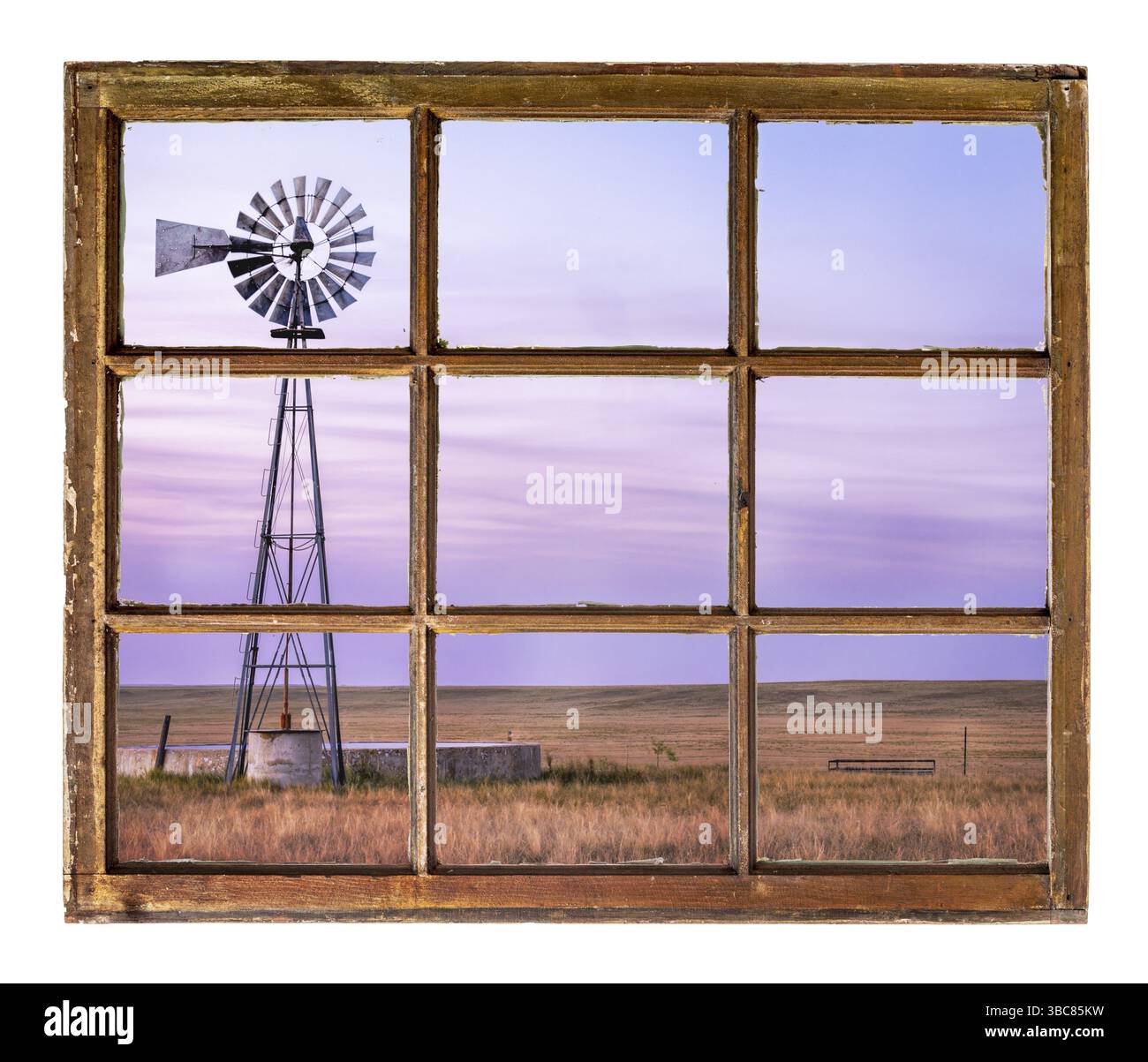 Mulino a vento con pompa e serbatoio d'acqua per bestiame nella prateria di erba corta al crepuscolo, visto dalla finestra di una cabina d'epoca Foto Stock