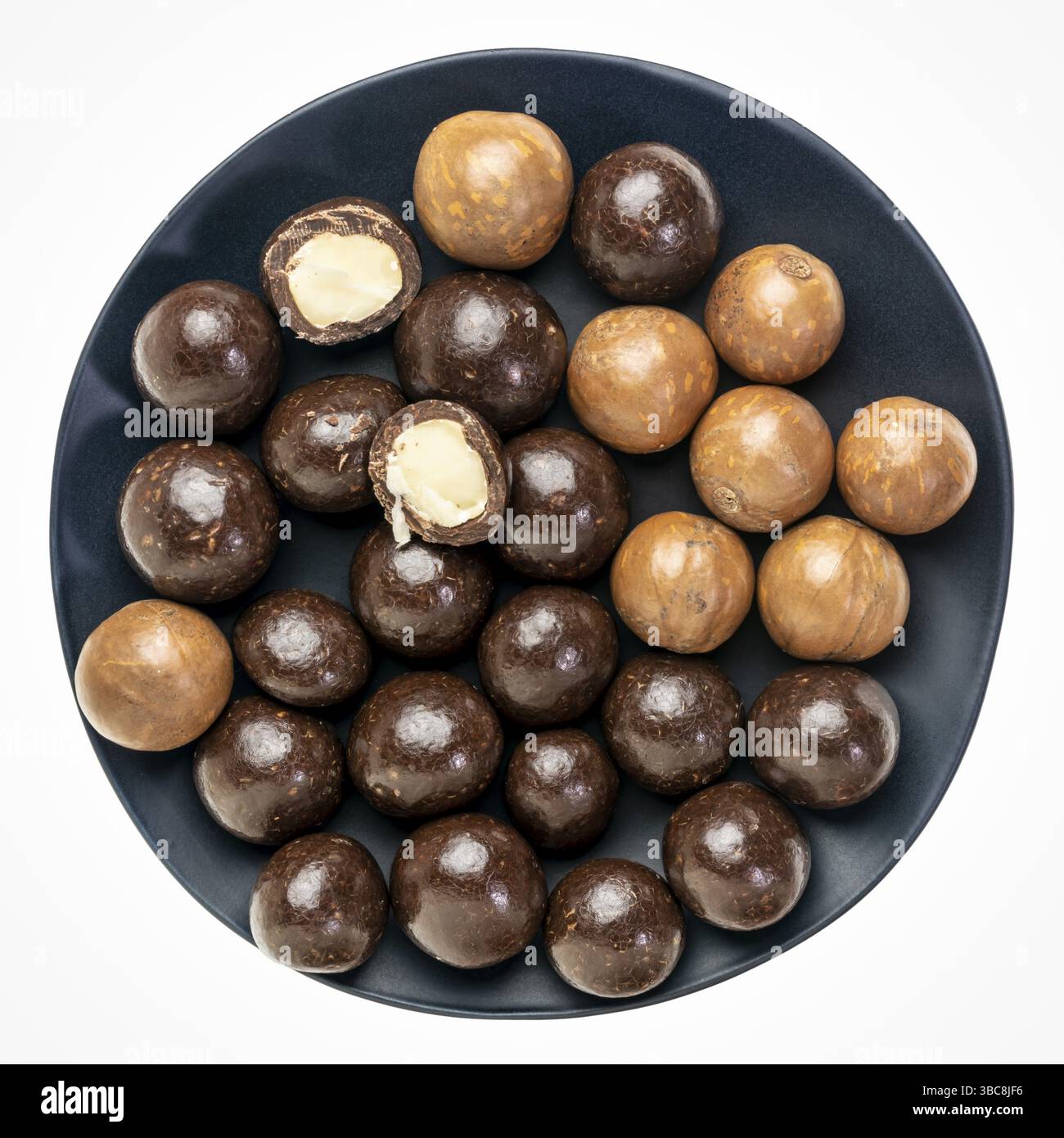 Noci di macadamia in conchiglie e immersi nel cioccolato fondente su un piatto nero isolato su bianco Foto Stock