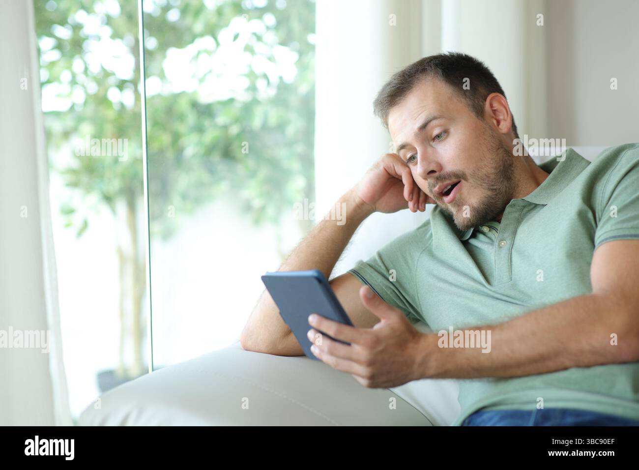 Uomo sorpreso che leggesse un eBook seduto su un divano in una casa al coperto Foto Stock