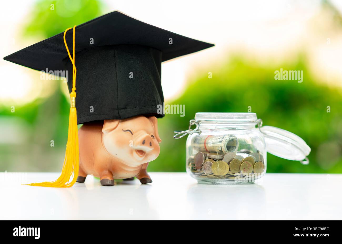 Un salvadanaio che indossa un cappello graduato con monete in un barattolo su sfondo sfocato. Foto Stock
