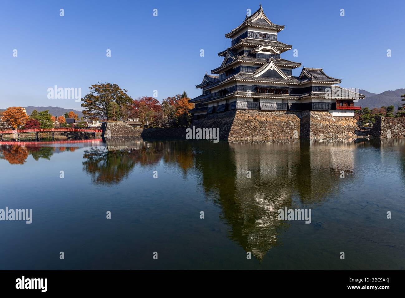 Vista panoramica del castello di Matsumoto con un ponte rosso, alberi d'autunno dorati e riflessi sull'acqua liscia in una mattina croccante Foto Stock