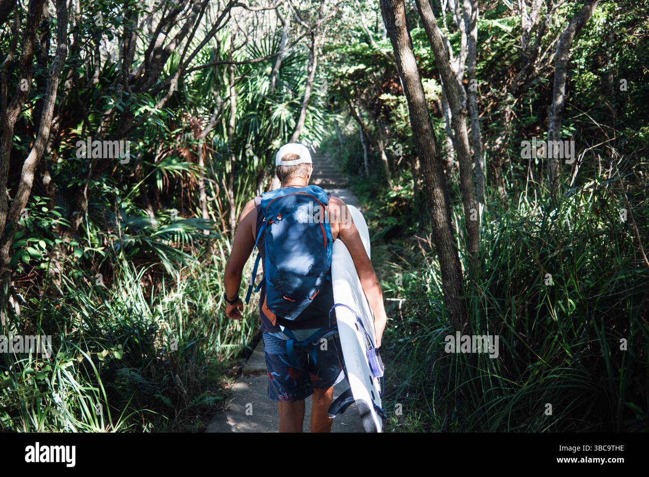 Il surfista cammina lungo il sentiero della foresta con tavola da surf e zaino Foto Stock