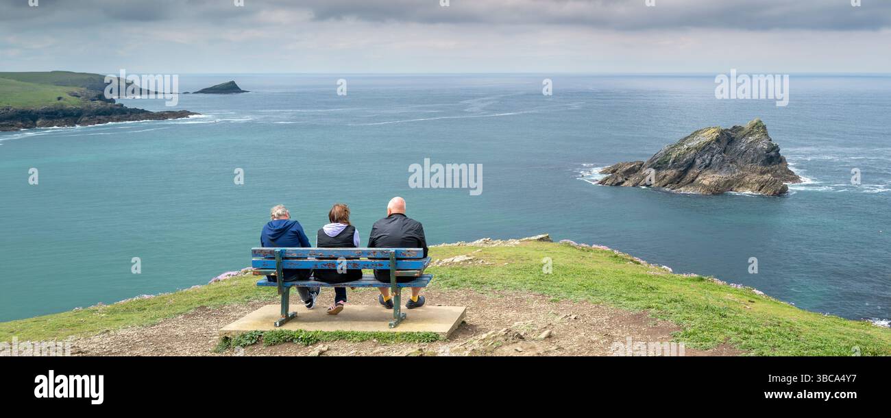 Una vista panoramica di tre turisti seduti su una panchina in cima a Pentire Point East che mostra le piccole isole rocciose, l'Oca Rock An Foto Stock