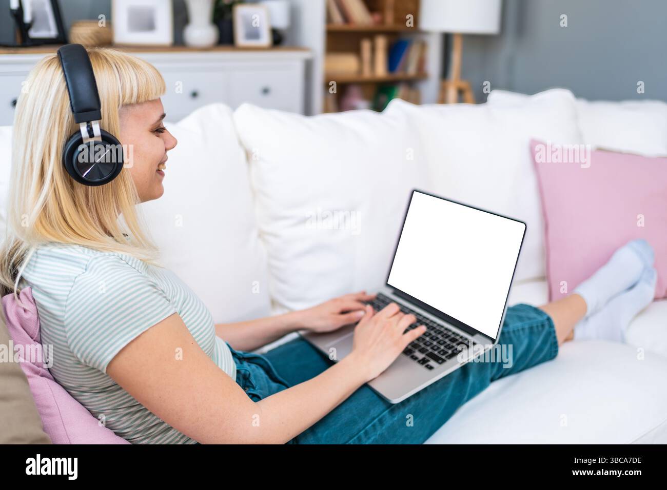Una giovane donna scambia lo studente seduto sul divano di casa utilizzando un computer portatile con cuffie wireless per lavorare in remoto partecipando a lezioni virtuali e riunioni video Foto Stock