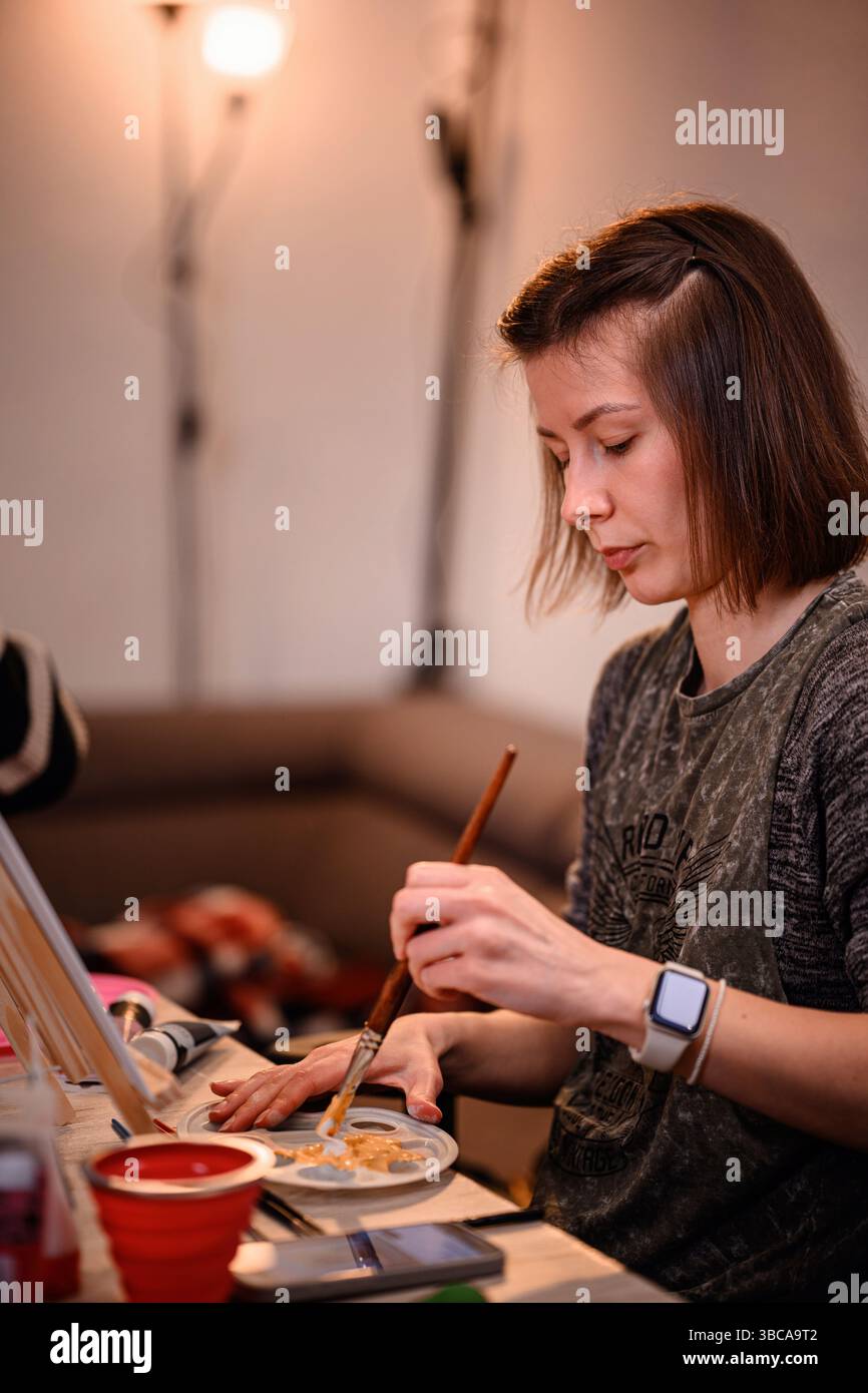 Una giovane donna incentrata fonde i colori di una tavolozza con un pennello, preparandosi a creare opere d'arte in un accogliente spazio interno. Foto Stock