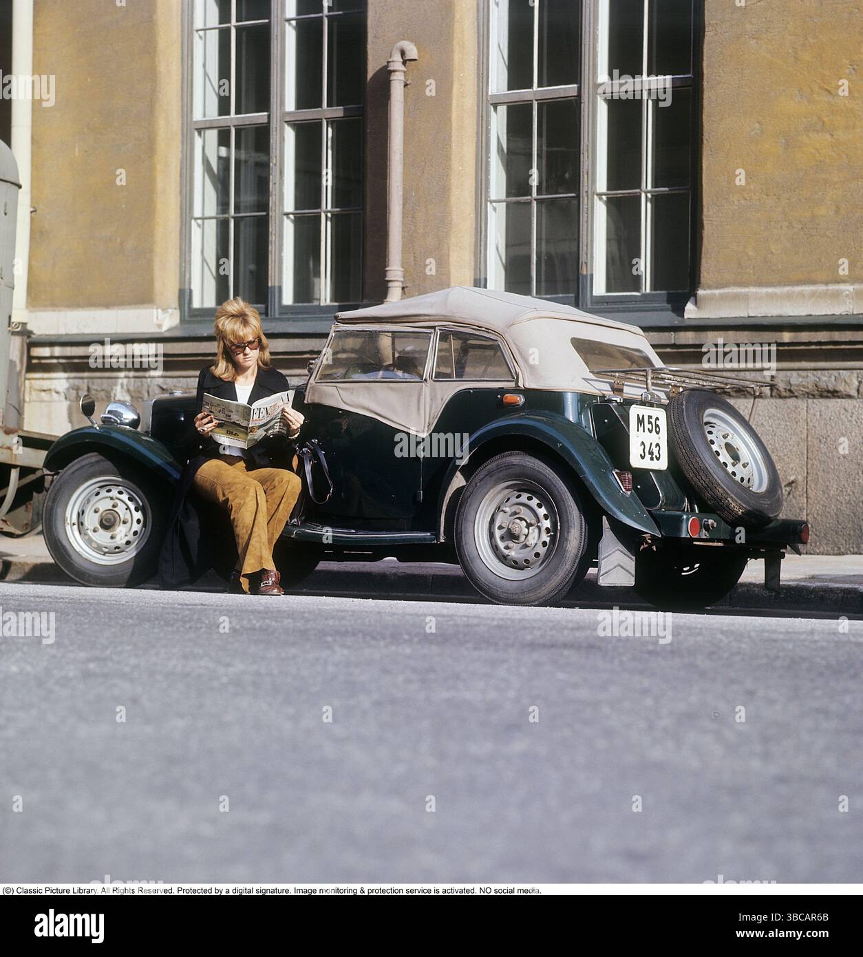 Una donna vicino a un'auto sportiva. La vettura è un modello MG TD inglese, prodotto tra il 1950 e il 1953. Questo modello era particolarmente adatto per i clienti svedesi in quanto poteva essere costruito con il volante sul lato sinistro, in linea con le convenzioni di guida svedesi dell'epoca. Sono state prodotte in totale 29.664 unità dell'MG TD. È seduta sulla macchina, a leggere la rivista femminile Femina. È vestita con un blazer e pantaloni marroni. Svezia degli anni '1960. Ulteriori informazioni sulla MG TD la MG TD, parte della serie MG T-Type, era una roadster a due posti rinomata per il suo classico design britannico, Feat Foto Stock