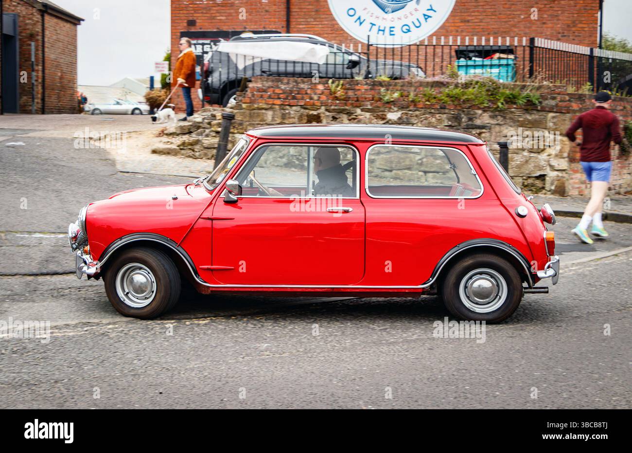 Una classica Mini Cooper rossa vibrante con tetto nero, che guida su una strada circondata da erba, con auto d'epoca ed edifici sullo sfondo. negli scudi nord Foto Stock