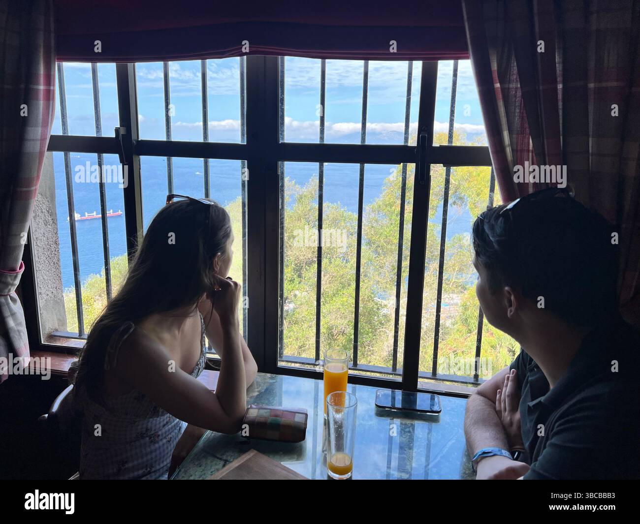Una coppia si siede vicino alla finestra di un caffè o ristorante sul mare, guardando le navi sull'oceano aperto e gli alberi lussureggianti sottostanti. Foto Stock