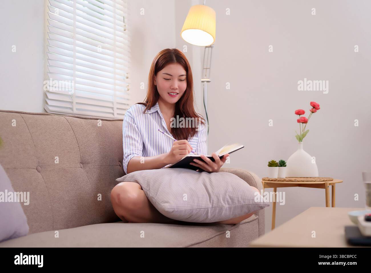Una giovane donna adulta è seduta su un divano a casa e scrive su un notebook. Sembra rilassata e concentrata, godendosi un momento di calma per riflettere in modo personale Foto Stock