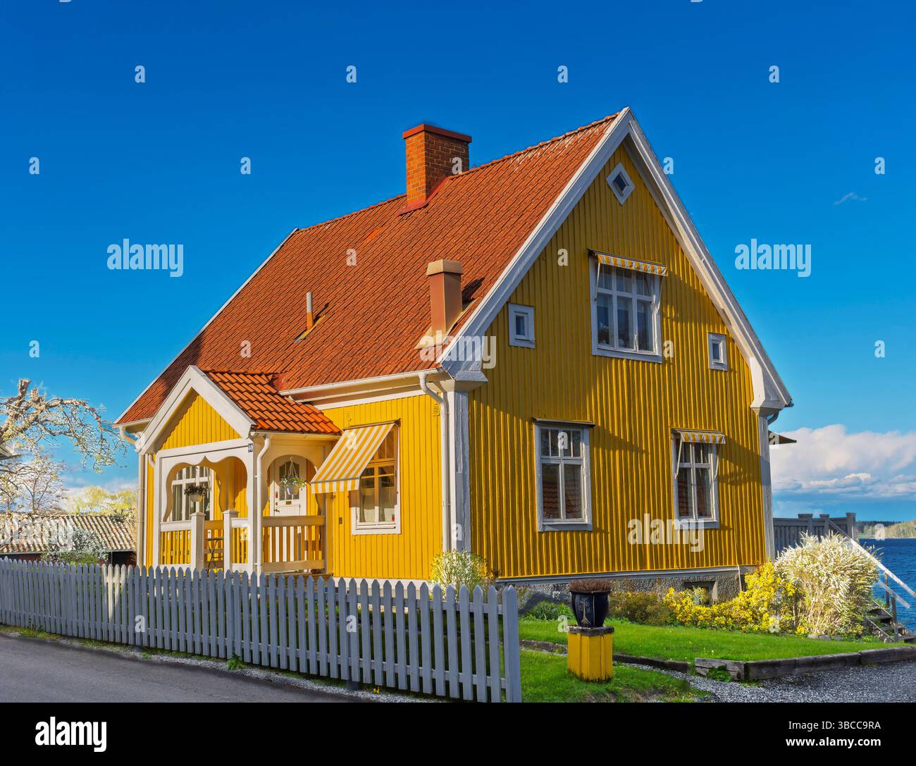 Tradizionale casa in legno giallo con tetto piastrellato rosso e recinzione a picchetto bianca sul lago in Svezia in una soleggiata giornata primaverile. Foto Stock