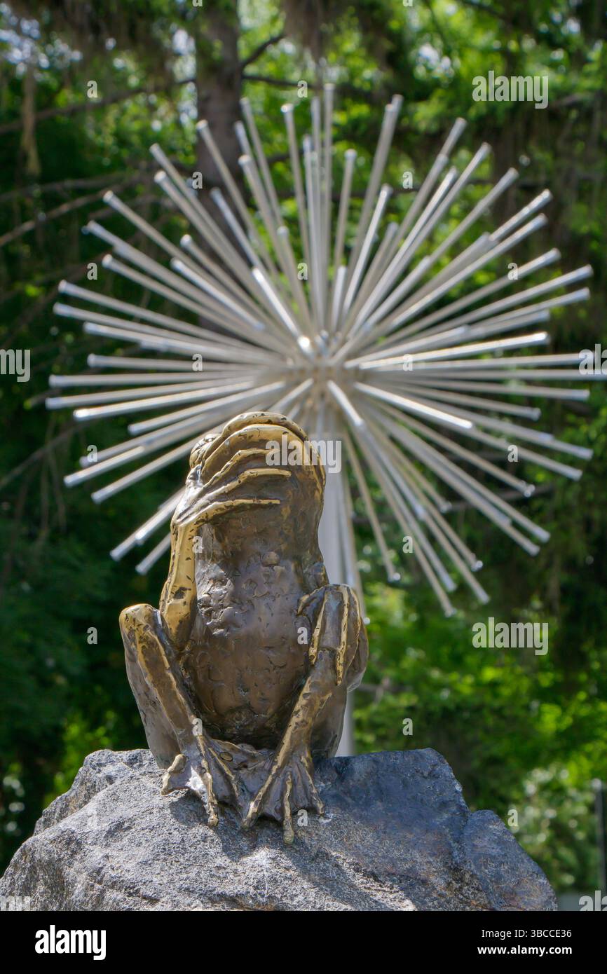 Una dettagliata scultura di rana si trova su una roccia in un lussureggiante giardino. Dietro di essa, il sorprendente design Sunburst in aste metalliche aggiunge un tocco moderno, il contrasto Foto Stock
