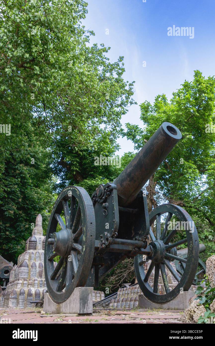 Un cannone storico si erge prominentemente in un parco, circondato da antiche strutture in pietra e da una vegetazione lussureggiante sotto un cielo azzurro. La scena cattura Foto Stock