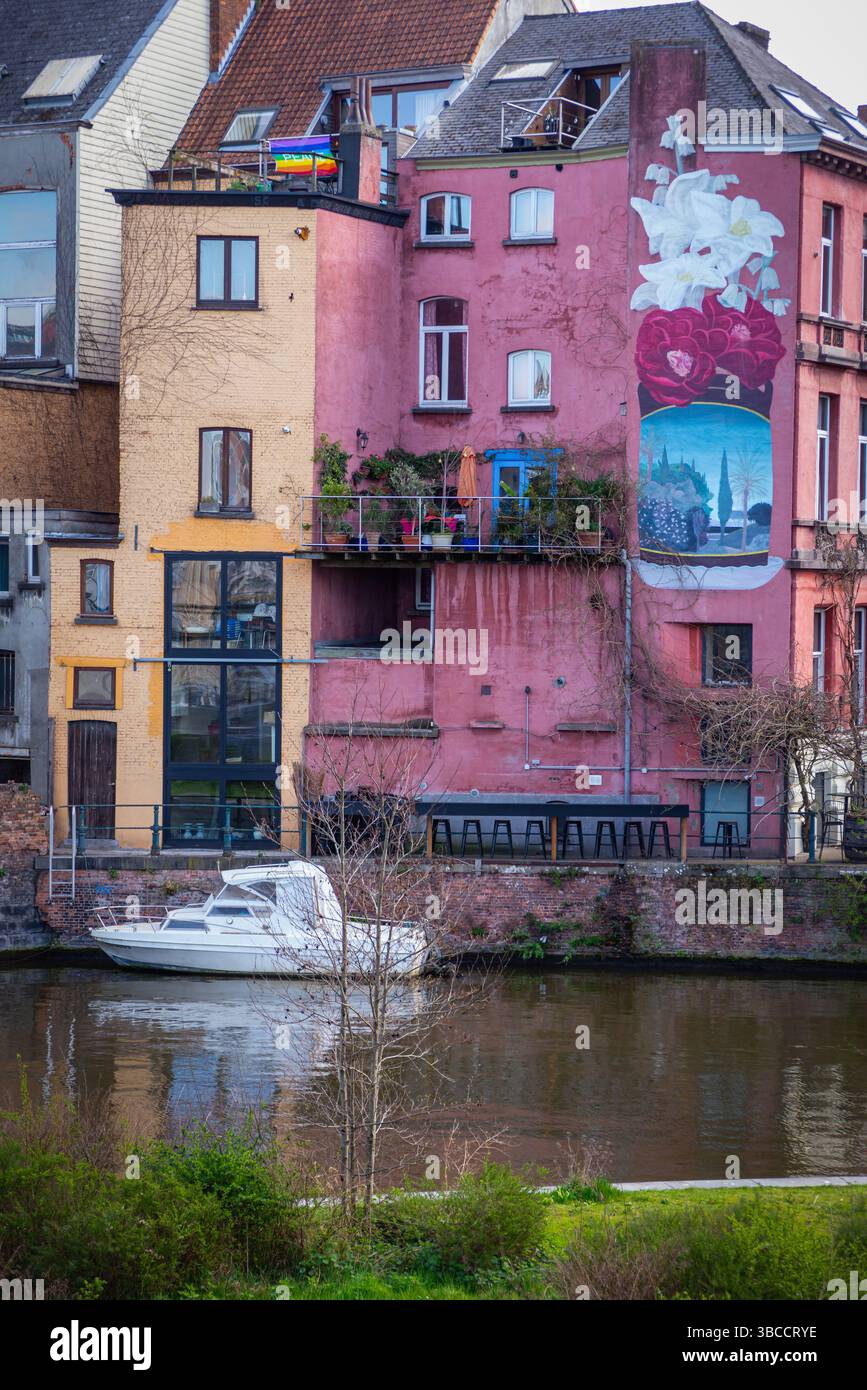 Una barca bianca galleggia su un canale a Gent, Belgio, di fronte a un edificio rosa lungo il fiume con un grande murale floreale, una bandiera arcobaleno, balconi, finestre, Foto Stock