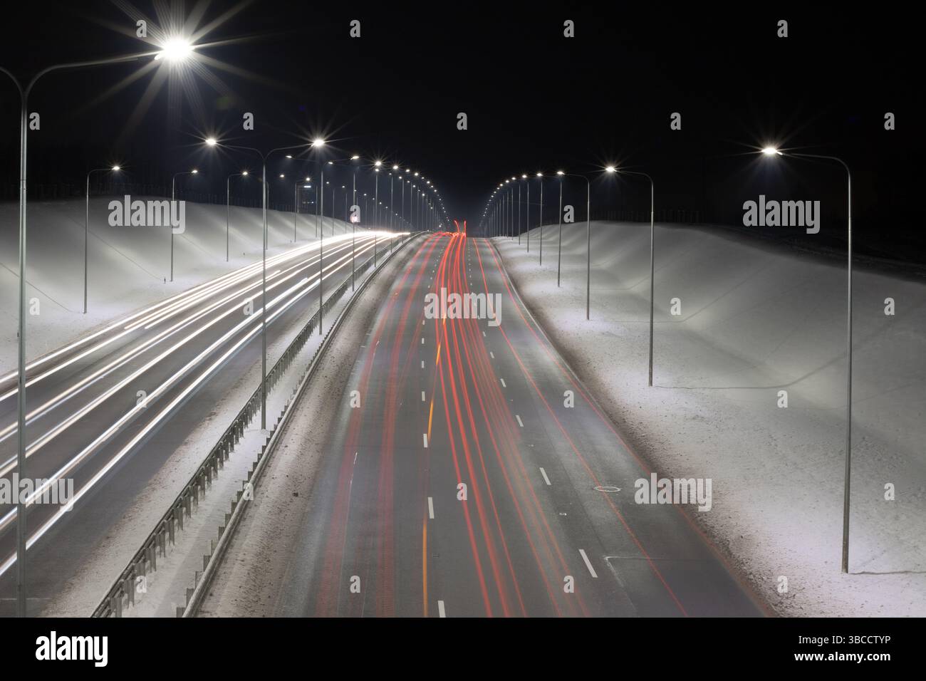 Una serena vista notturna di un'autostrada a più corsie incorniciata da terrapieni innevati e lampioni luminosi. Acquisizione con esposizione lunga Foto Stock