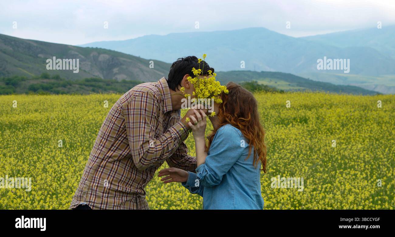 Giovane coppia che si baciano dietro un bouquet di fiori selvatici gialli in un fiorente campo primaverile. Momenti divertenti e romantici nella natura. Ideale per l'amore, lo stile di vita Foto Stock