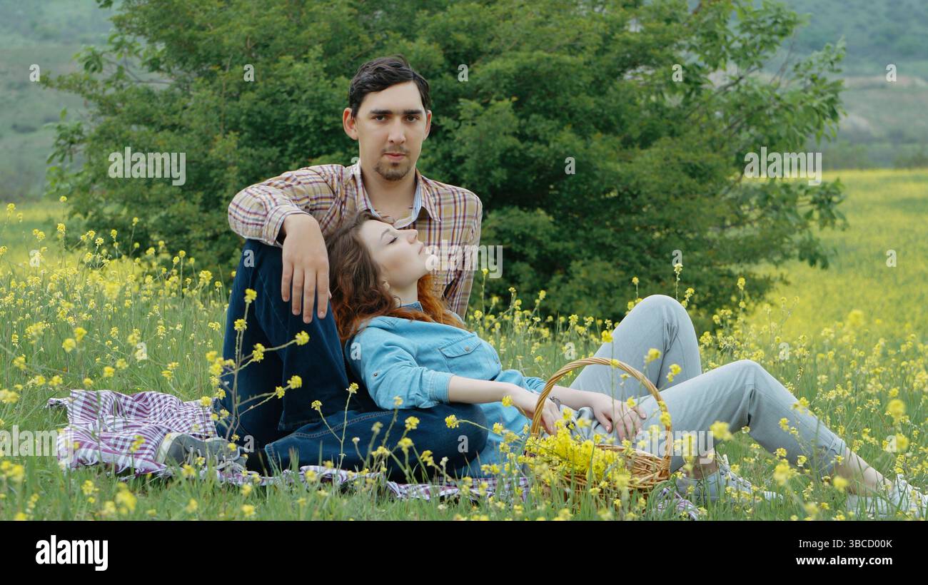 Coppia romantica che si gode un tranquillo picnic primaverile in un fiorito campo di fiori. La donna riposa sulle ginocchia dell'uomo, circondata da fiori selvatici gialli Foto Stock