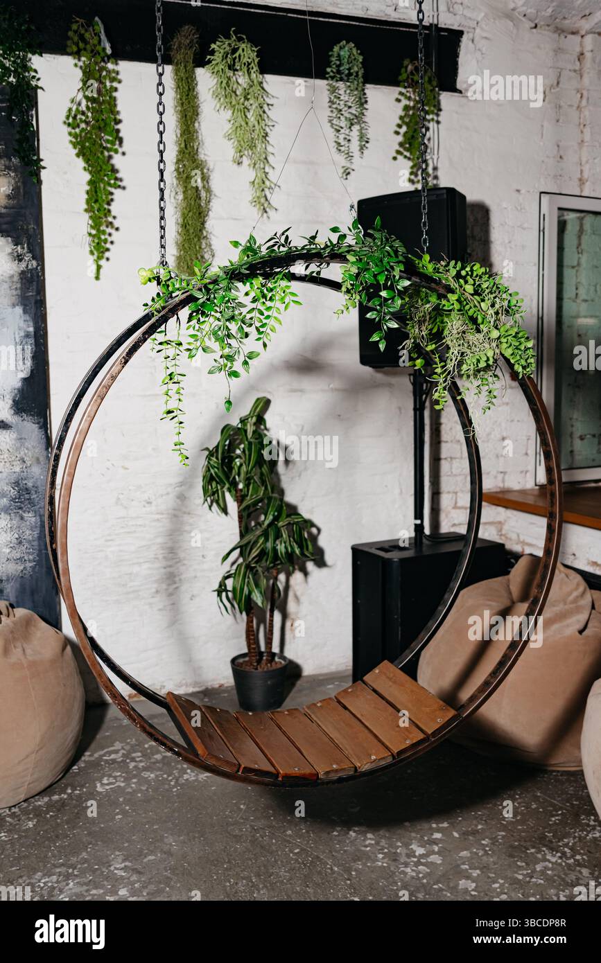 Elegante sedia a dondolo circolare in legno adornata con piante verdi, collocata in un accogliente e moderno loft interno con pareti in mattoni bianchi. Foto Stock
