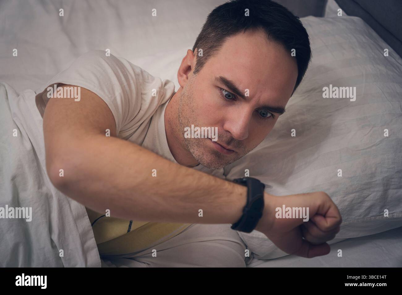 La testa di un giovane adulto sdraiata su un cuscino bianco nel letto. Hand consente di spegnere o accendere la sveglia. Non dormi abbastanza, svegliati. il tizio guarda la s. Foto Stock