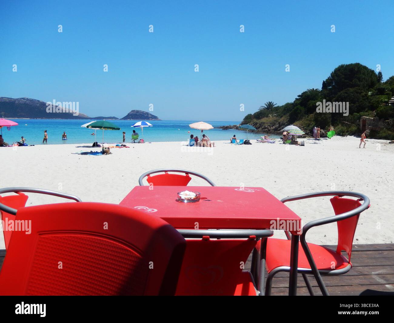 Sedie con tavolo in un bar sulla spiaggia nella baia di Golfo Aranci, Sardegna. Spiaggia di sabbia bianca di fronte a un mare azzurro brillante,Golfo Aranci, Sardegna, Italia Foto Stock