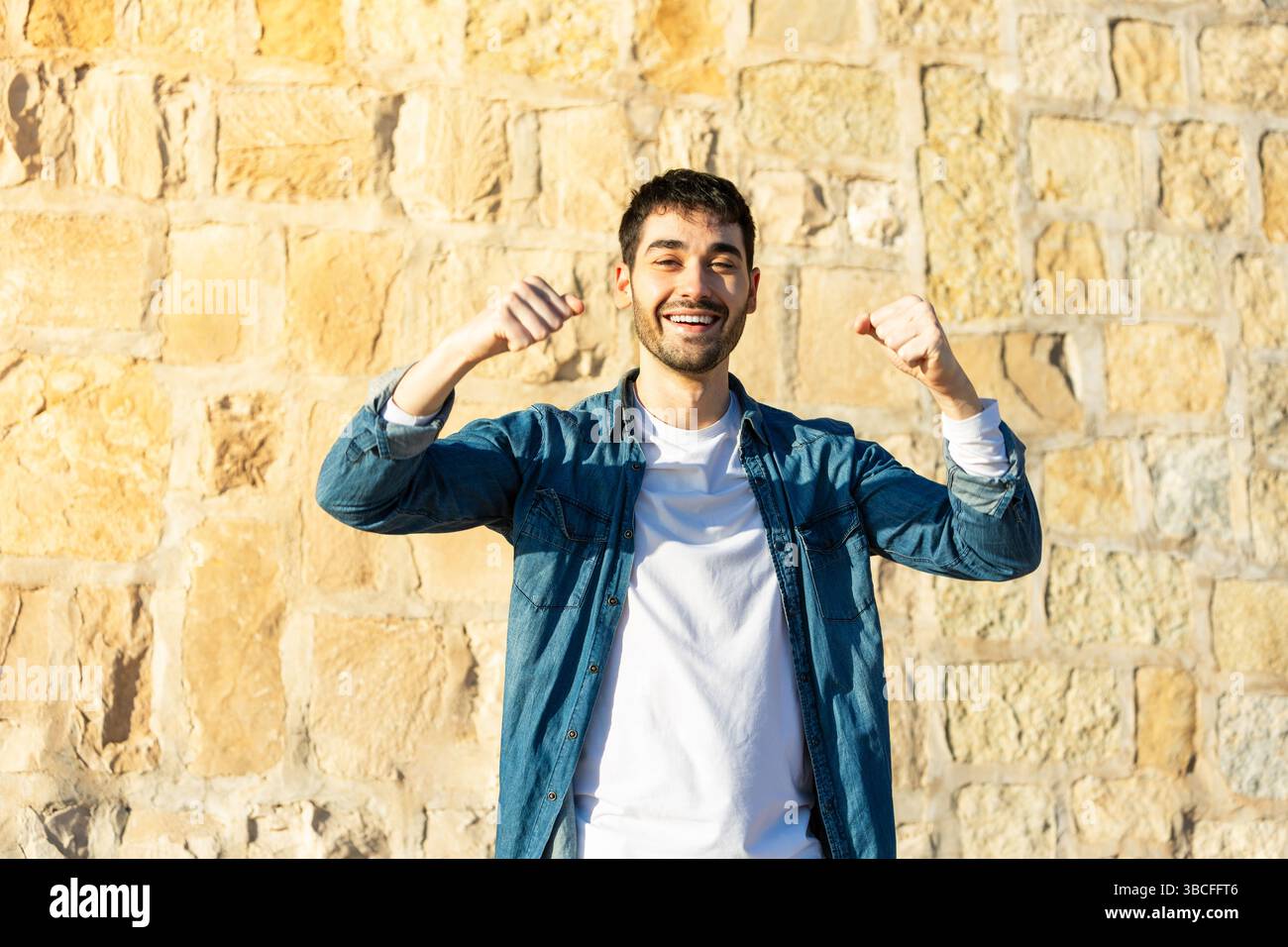 Un giovane sorridente in una camicia casual in denim che solleva entrambi i pugni per festeggiare mentre si trova all'aperto su uno sfondo di pietra illuminato dal sole. Stile di vita. Foto Stock