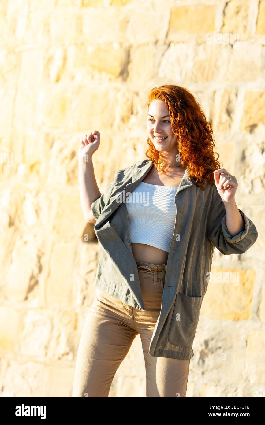 Gioiosa donna dai capelli rossi con un grande sorriso che danza casualmente alla luce del sole, braccia sollevate e piene di energia, in piedi contro un muro di pietra in un outfit casual. Foto Stock