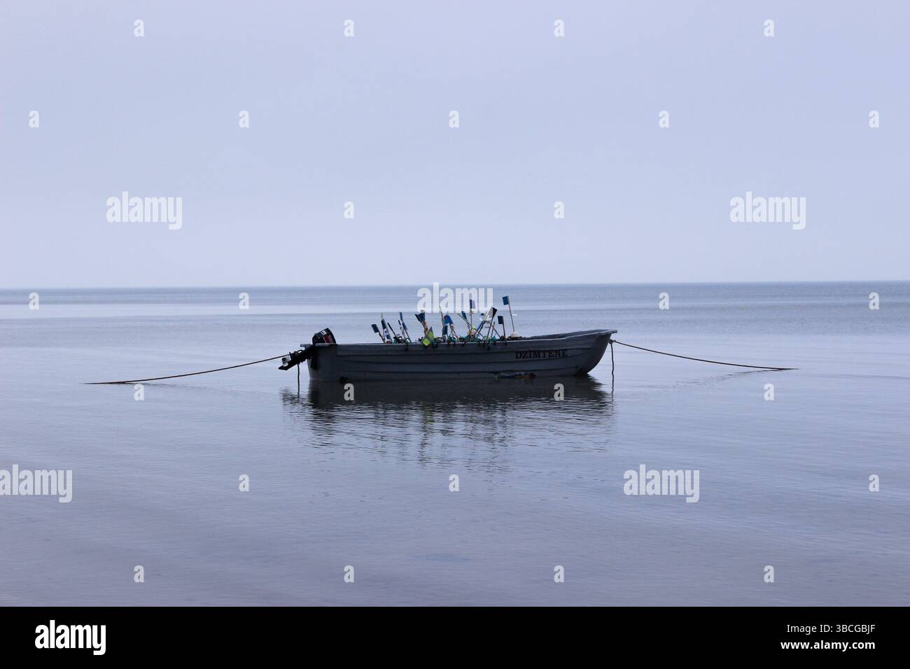 Una barca da pesca solitaria chiamata “DZIMTENE” ancorata nelle acque calme e riflettenti di un mare sotto un cielo coperto. Foto Stock