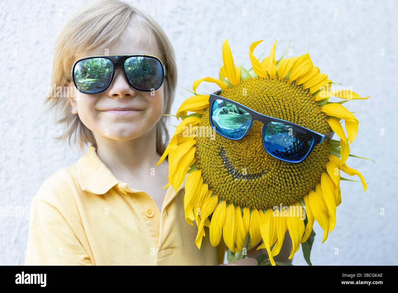simpatico bambino di 7 anni con occhiali da sole e fiori di girasole con un sorriso tagliato. Somiglianza delle immagini. Atmosfera estiva, fantasie floreali, atmosfera positiva. Foto Stock