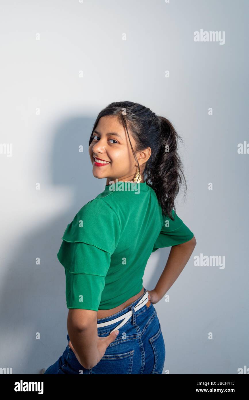 Una donna con una camicia verde e un jeans blu sorride per la macchina fotografica. Indossa un orecchino d'oro e una collana d'oro Foto Stock Una donna con una camicia verde e un jeans blu sorride per la macchina fotografica. Indossa un orecchino d'oro e una collana d'oro Foto Stock