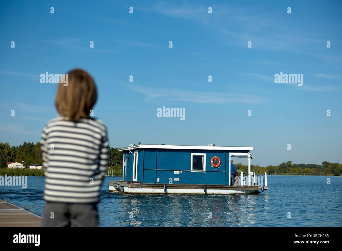 Un bambino in camicia a righe guarda una casa galleggiante blu su un lago calmo sotto un cielo limpido. Altopiano del lago di Meclemburgo, Meclemburgo-Pomerania occidentale, Germania Foto Stock