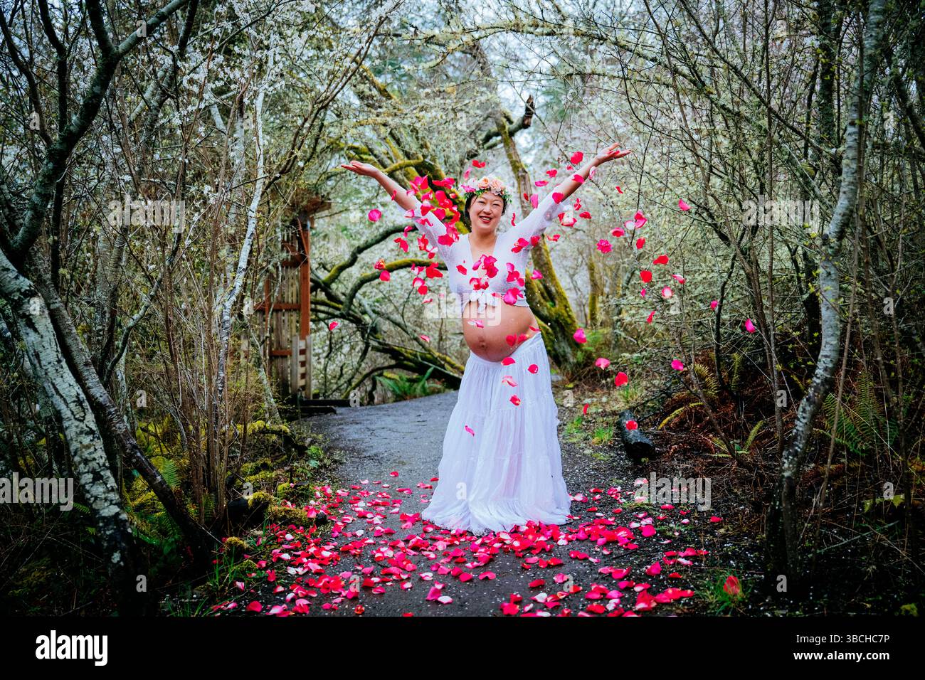 Donna incinta in abito bianco che festeggia nella foresta circondata da petali rosa. WASHINGTON, STATI UNITI Foto Stock