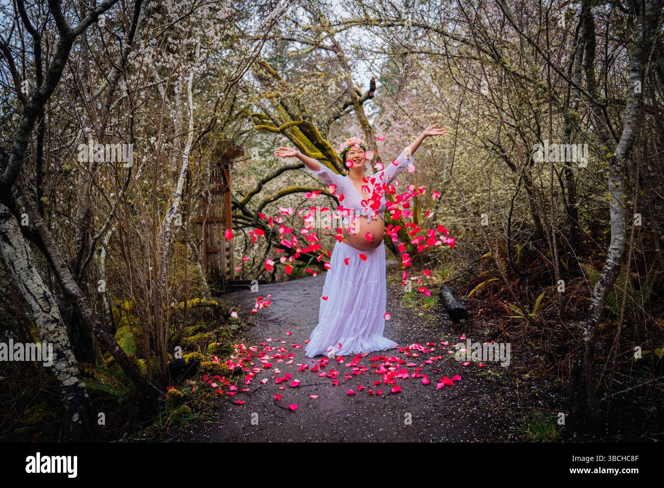 Donna incinta in abito bianco che festeggia nella foresta circondata da petali rosa. WASHINGTON, STATI UNITI Foto Stock