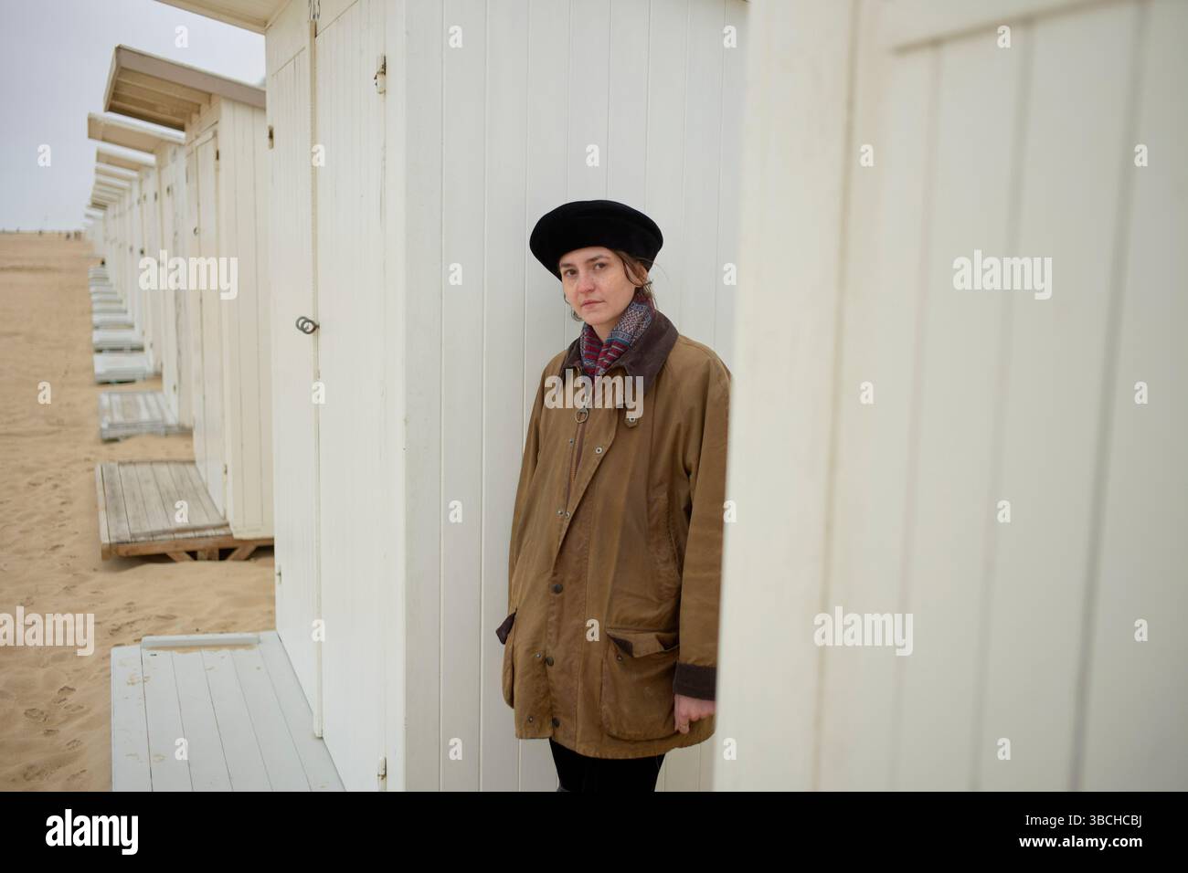 Donna con un cappotto marrone e berretto in piedi accanto a capanne bianche sulla spiaggia di sabbia. Ostenda, Belgio Foto Stock