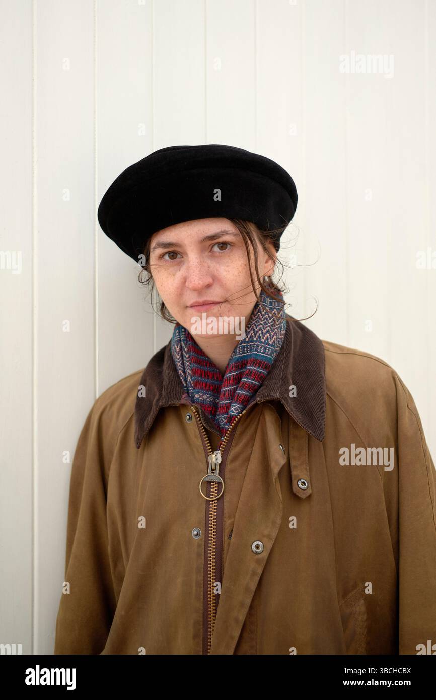 Una donna con un cappotto e un cappello marroni si staglia accanto a una parete di legno bianca, guardando con sicurezza la macchina fotografica. Ostenda, Belgio Foto Stock
