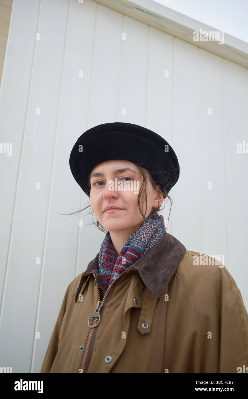 Una donna con un cappotto e un cappello marroni si staglia accanto a una parete di legno bianca, guardando con sicurezza la macchina fotografica. Ostenda, Belgio Foto Stock
