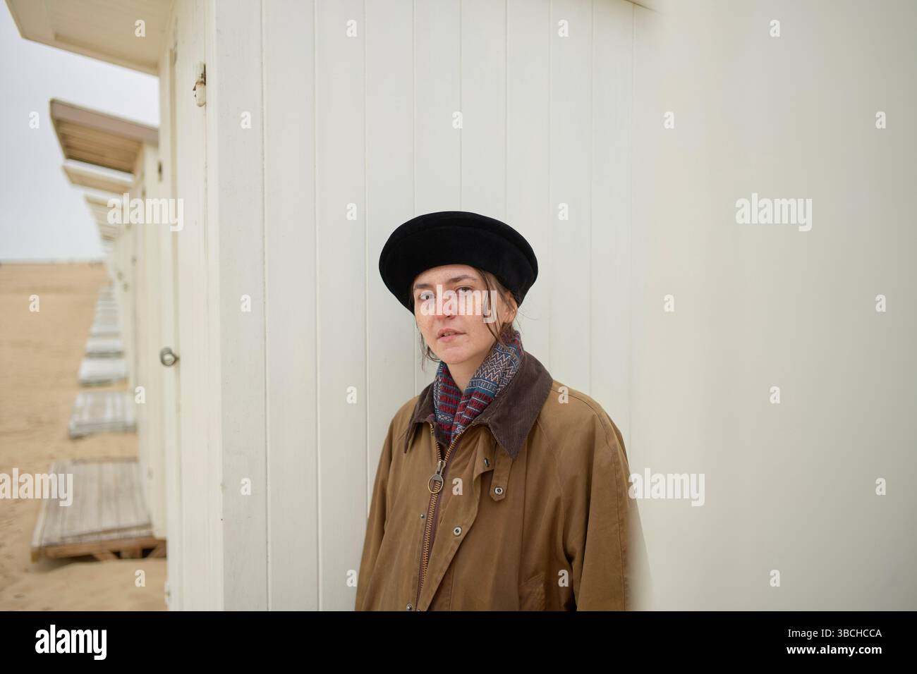 Donna con un cappotto marrone e berretto in piedi accanto a capanne bianche sulla spiaggia di sabbia. Ostenda, Belgio Foto Stock