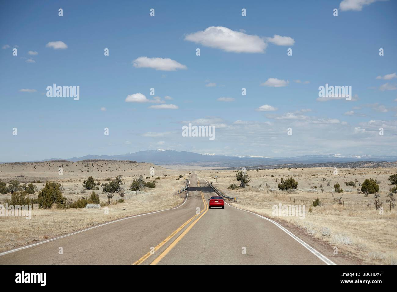 Un'auto rossa percorre una lunga e vuota strada attraverso un vasto e asciutto paesaggio sotto un cielo azzurro. STATI UNITI Foto Stock