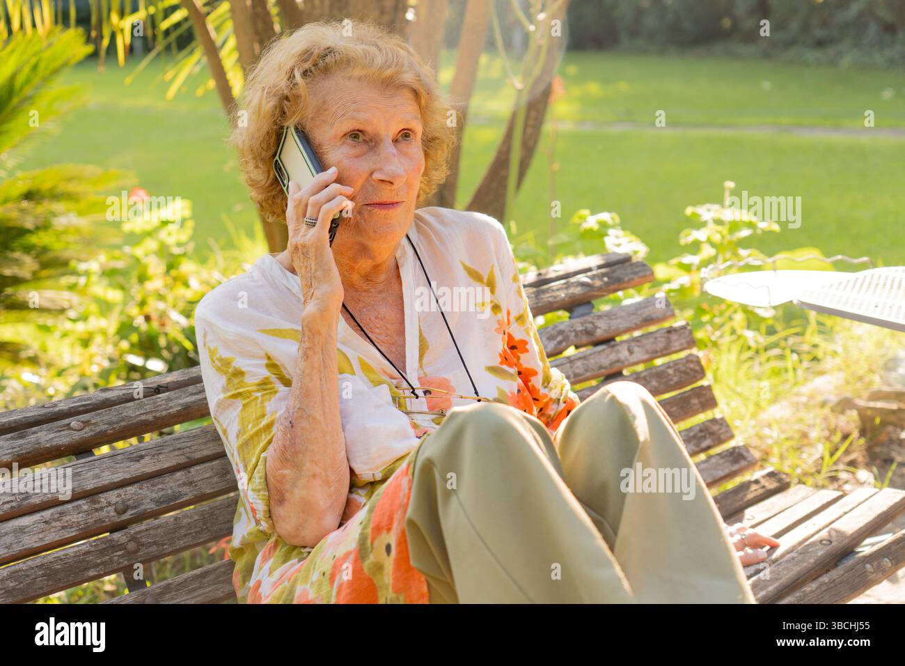 Donna anziana che si gode una conversazione sul cellulare mentre siede su una panchina di legno in un tranquillo parco, abbraccia la tecnologia e resta co Foto Stock