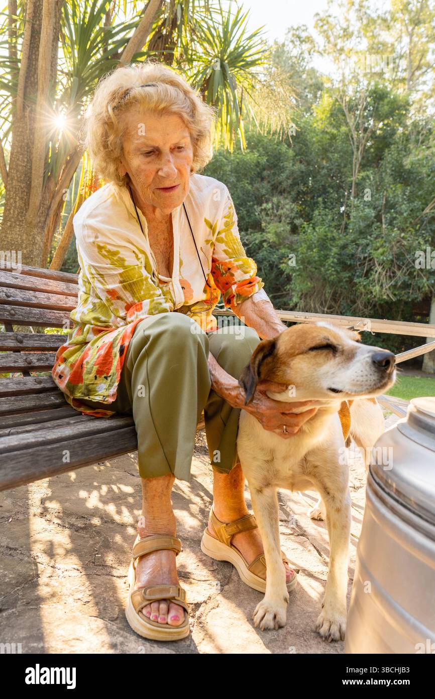 Donna anziana che si gode un momento di pace all'aperto, seduta su una panchina di legno nel suo cortile e accarezzare affettuosamente il suo cane sotto la calda luce del sole Foto Stock