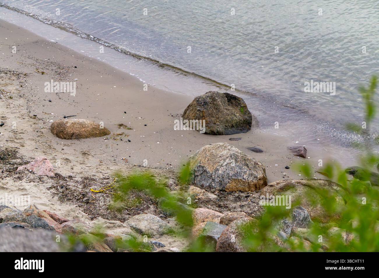 Spiaggia sabbiosa con rocce e vegetazione in primo piano, dolci onde che bagnano la riva. Paesaggio costiero naturale dai toni tenui. Foto Stock