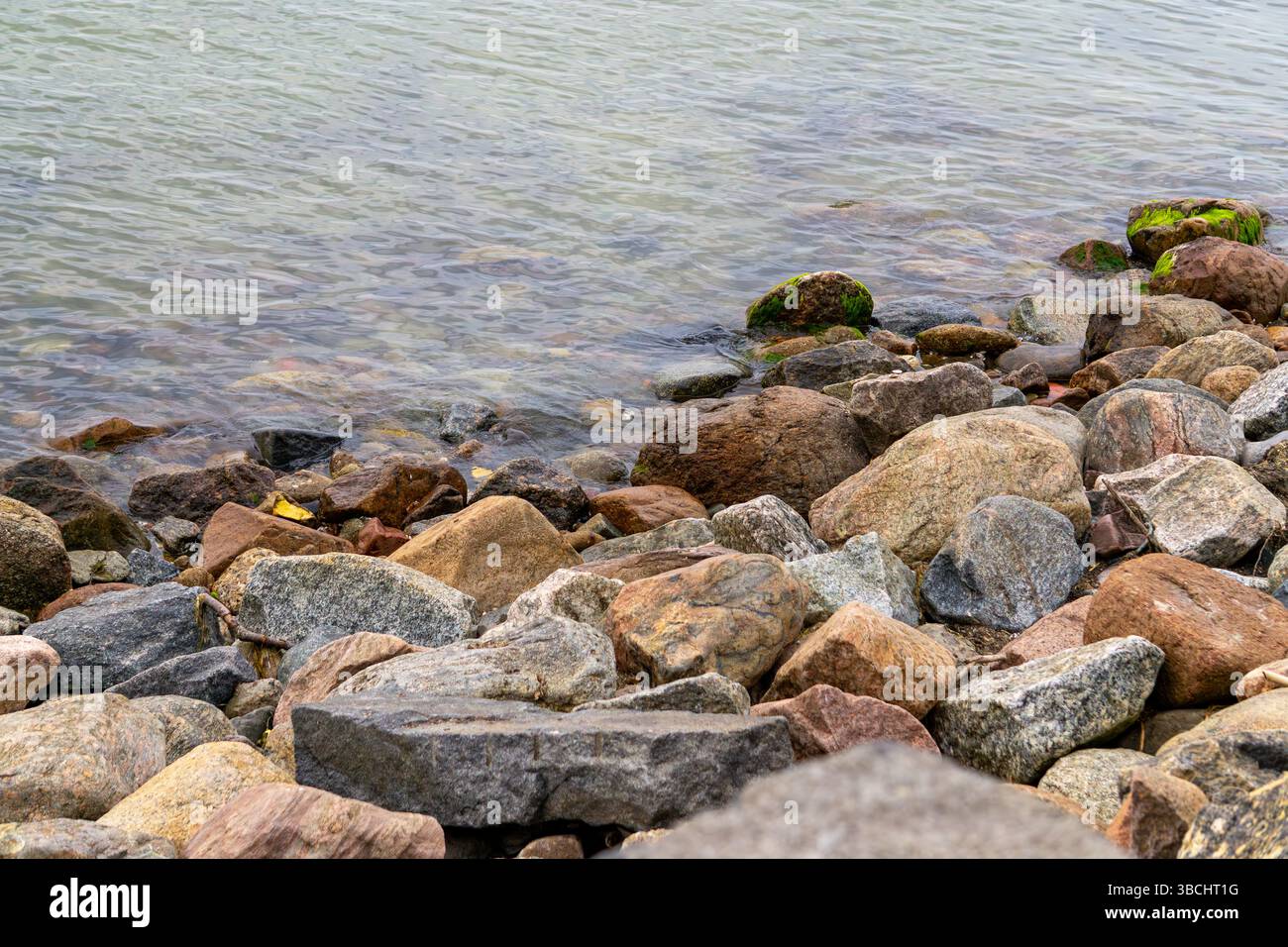 Spiaggia sabbiosa con rocce e vegetazione in primo piano, dolci onde che bagnano la riva. Paesaggio costiero naturale dai toni tenui. Foto Stock