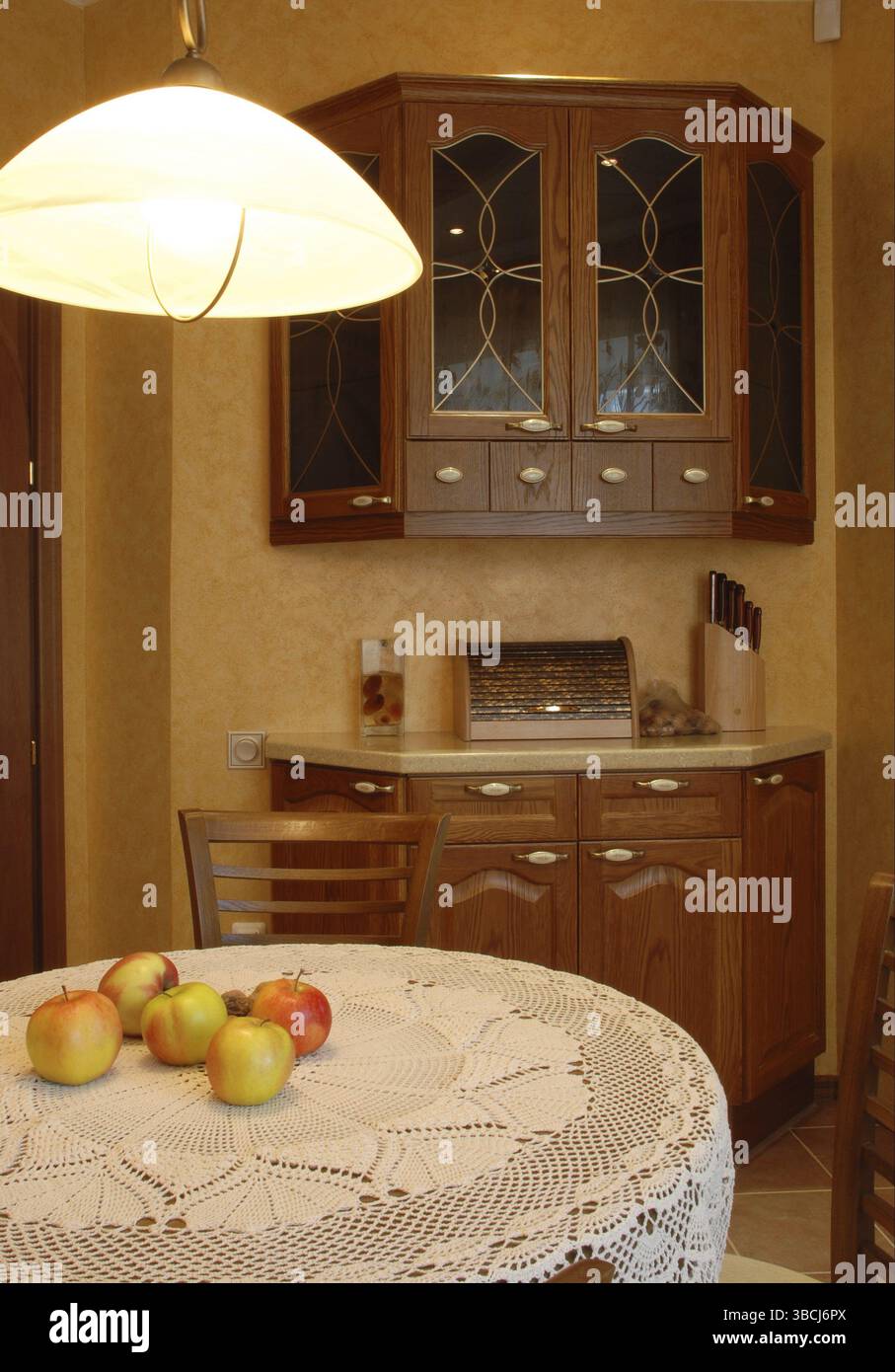 Cucina accogliente: Armadietti in noce, tavolo rotondo ricoperto da un bellissimo tovagliolo bianco all'uncinetto con mele rubicund su di esso, lampada abbassata Foto Stock