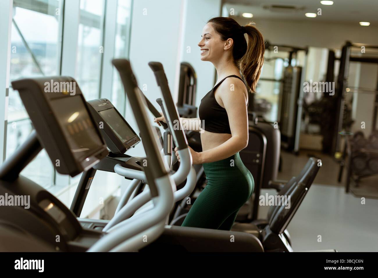 L'appassionato di fitness ama allenarsi sull'ellittica in una palestra luminosa e ariosa piena di luce solare e attrezzature, dimostrando la sua dedizione alla salute. Foto Stock