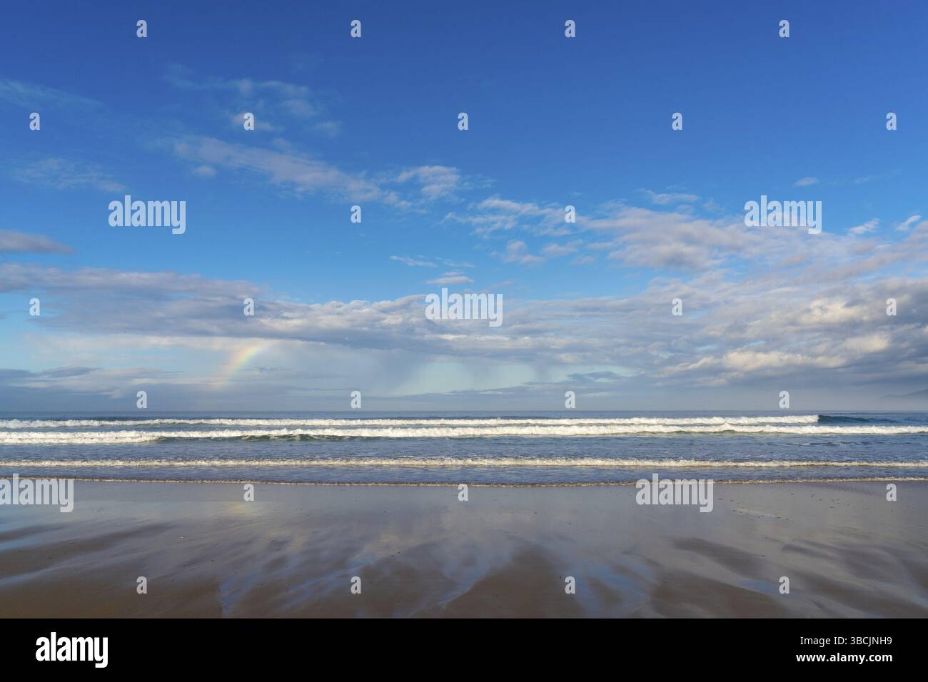 Una spiaggia di sabbia con grandi onde e un piccolo arcobaleno in un cielo espressivo Foto Stock