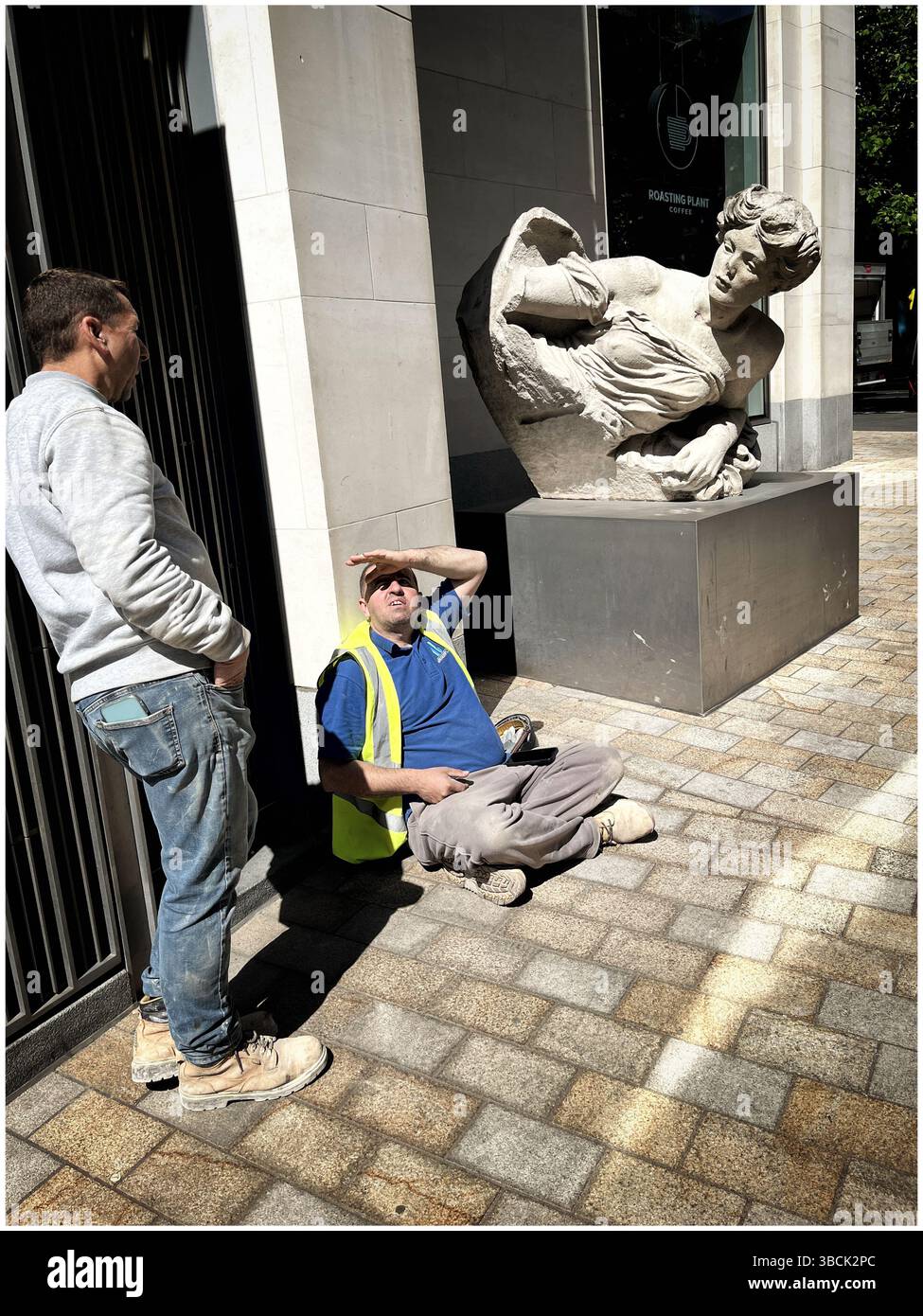 Due operai in una strada di Londra, accanto a una statua, che si rompono, uno che protegge gli occhi dal sole. Foto Stock