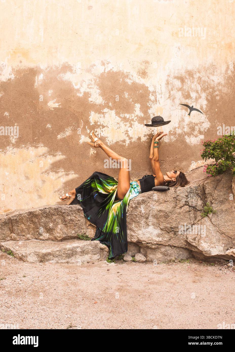 Una donna glamour si stende con un cappello e si rilassa accanto al muro di pietra, mentre mostra la sua lunga gamba fuori dalla gonna di seta. La modella si sta divertendo. Foto Stock