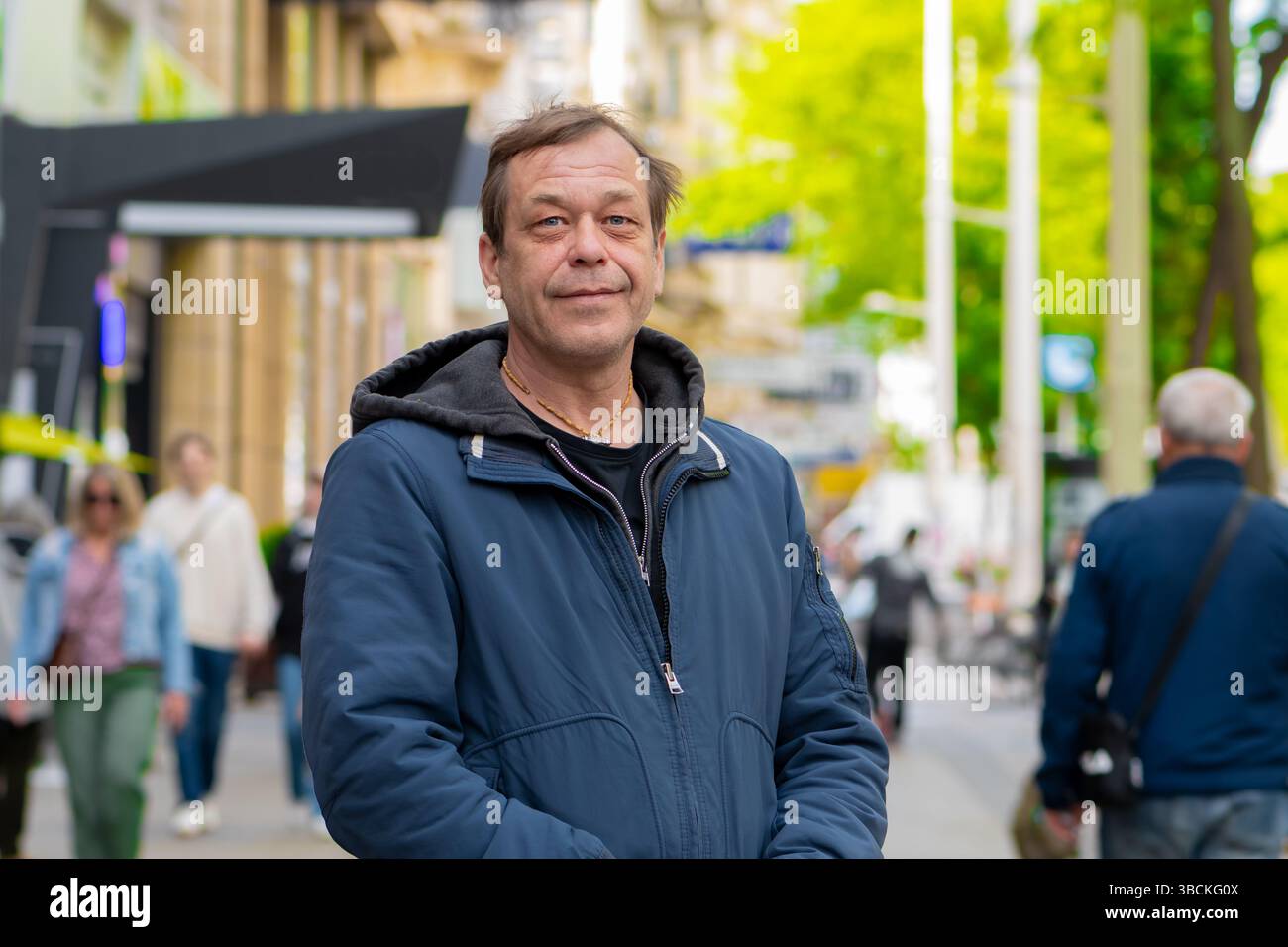 Ritratto di un uomo adulto di 50-55 anni con una giacca blu in piedi su una strada trafficata della città, su uno sfondo sfocato di passanti ed edifici Foto Stock