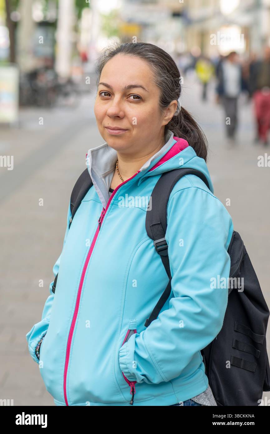 Donna di 40-45 anni con una giacca sportiva blu con zaino in piedi su una strada della città - stile casual e stile di vita attivo Foto Stock