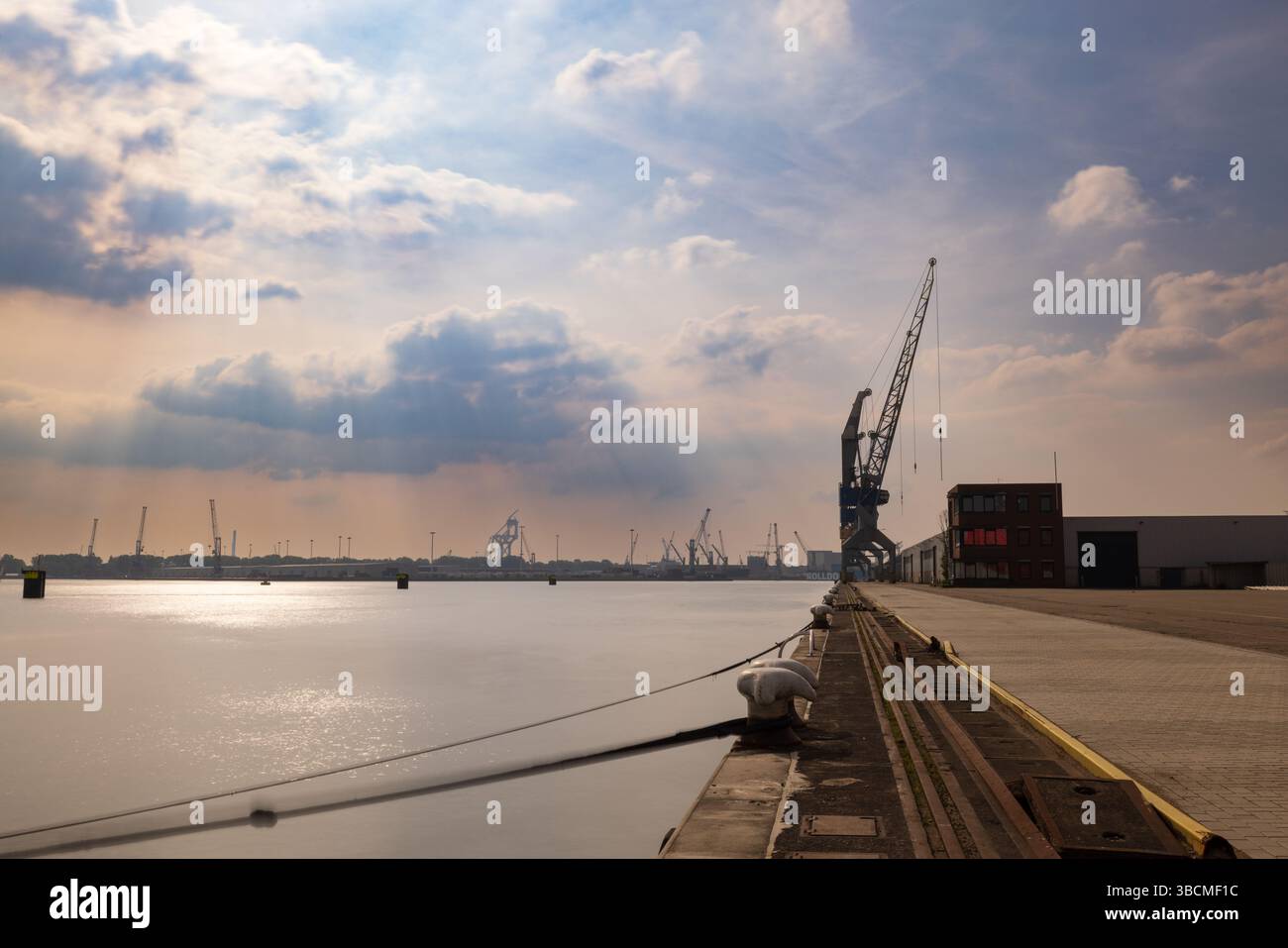 Molo industriale con gru lungo il porto di Rotterdam, inondato di luce calda sotto una spettacolare copertura nuvolosa e acque calme. Tecnica di esposizione lunga. Foto Stock