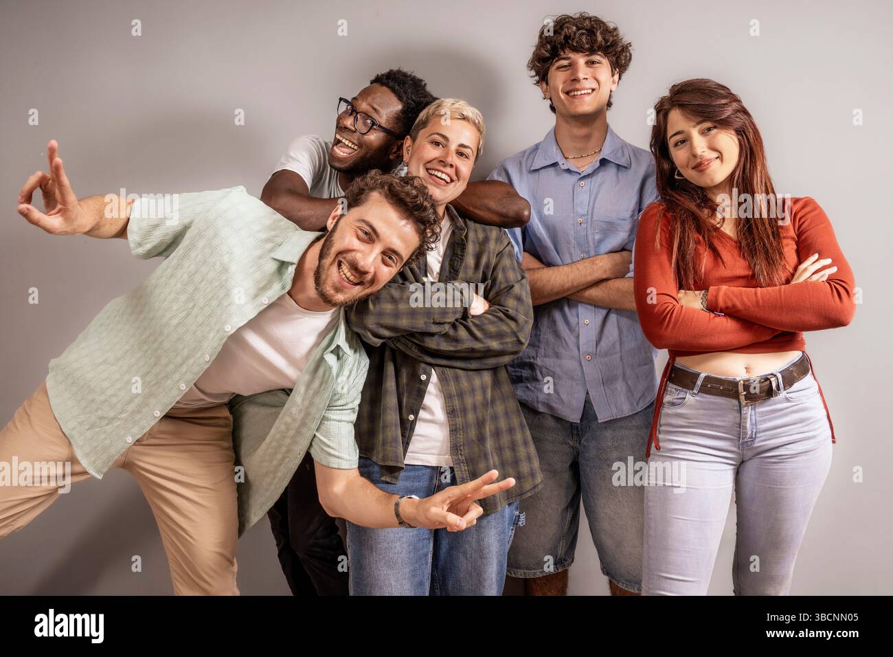 Cinque giovani adulti multiculturali ridono e posano insieme in un luminoso studio, celebrando l'amicizia e l'unità in un ritratto di gruppo informale e sincero. Foto Stock